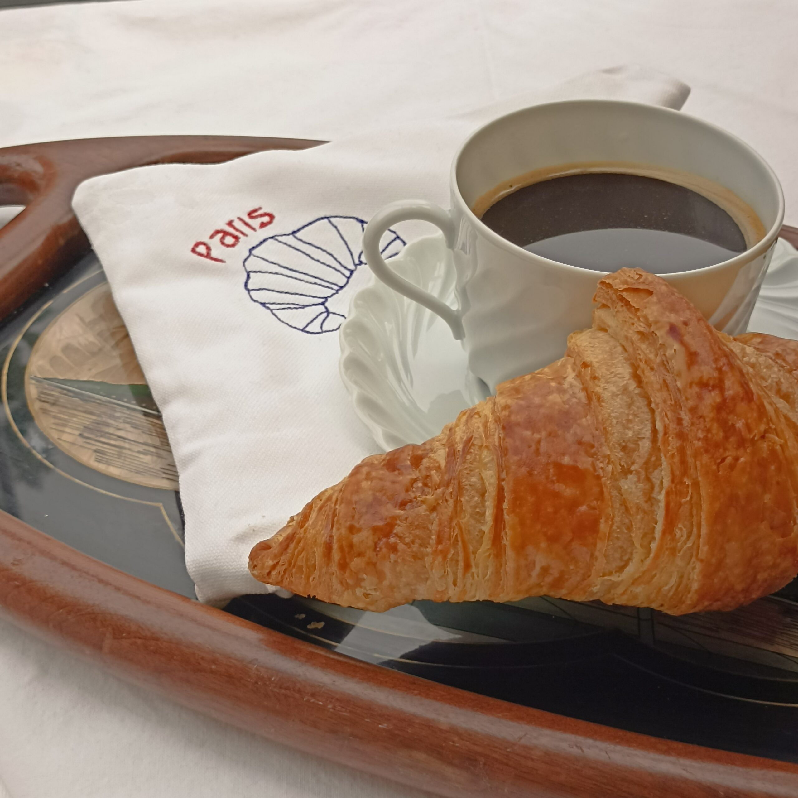 Serviette brodée « Paris » – Création de l’ESAT de la Bièvre Serviette de table en coton brodée d’un croissant et de l’inscription « Paris », présentée avec une tasse de café et un croissant.
